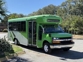 A green and white 2013 Chevrolet Express bus with large windows and a raised roof is parked with its doors open