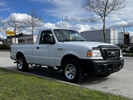 A white 2010 Ford Ranger pickup truck with a cab and bed parked on the street reflecting sunlight on its shiny chrome wheels