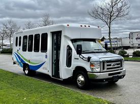 A white 2018 Ford Econoline shuttle van with blue and green graphics is parked, featuring an open door and accessible ramp