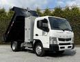 A 2015 Mitsubishi Fuso FE dump truck with a white cab and a gray metal storage compartment, featuring a raised black dump bed and orange marker lights on the roof