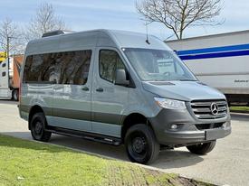 A 2019 Mercedes-Benz Sprinter van in a sleek gray color with large windows and a streamlined design parked on the side of the road