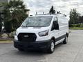 A white 2023 Ford Transit van with a roof rack and black front grille parked on a concrete surface