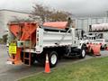 A 2012 International 7400 snow plow truck with a large orange plow attached to the front and a white cargo box in the back displaying a caution sign