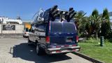 A blue 2013 Ford Econoline van with a ladder rack on top and large speakers mounted at the rear