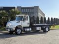 A 2017 International 4300 flatbed truck with a white cab and black flatbed parked on a street with palm trees nearby