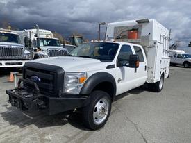 A white 2015 Ford F-550 truck with a flatbed and utility box on the back, featuring an armored grille and orange lights on top