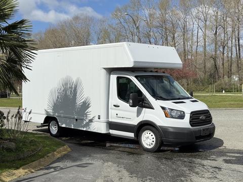A 2016 Ford Transit cargo van with a white boxy cargo area and a black front grille parked on a driveway