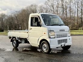 A white 2008 Suzuki Carry pickup truck with a flatbed and a compact design parked on a wet surface