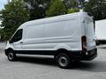 A white 2022 Ford Transit van with a high roof and sliding side door parked on a surface with trees in the background