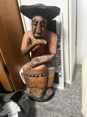A rare wooden sculpture of a seated figure playing a drum wearing a hat with intricate carvings on its body and facial features