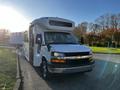 A 2017 Chevrolet Express bus with a white exterior and logo on the front, featuring large windows and side mirrors, parked with the front facing slightly to the left