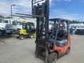 A 2010 Toyota 7FGGU25 forklift with a black and orange color scheme has raised forks and is positioned in the foreground with a seating area for the operator
