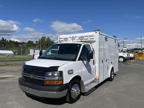 A white 2016 Chevrolet Express with a utility service body and a black front grille parked in a lot with silver wheels and emergency lights on the roof