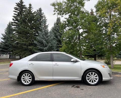 A silver 2012 Toyota Camry parked on a paved area with trees in the background. The car is shown from the side with tinted windows and alloy wheels