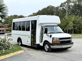A white 2015 Chevrolet Express shuttle bus with large windows and an open door for passenger access