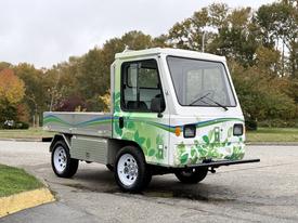 A 2019 CanEV Might-E electric utility vehicle with a white body and green leaf designs on the sides features a cab with large windows and a flatbed for cargo.