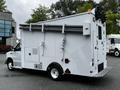 A white 2013 Ford Econoline van with a boxy cargo area and large pipes mounted on the roof equipped with various external compartments and lights