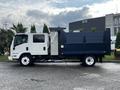 A 2015 Isuzu NPR HD truck with a white cab and a dark blue dump bed parked on a concrete surface