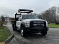 A 2015 Ford F-550 with a flatbed and tool storage on top features a black front grille and multiple orange safety lights on the roof