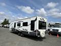 A white and gray 2013 Outdoor Wind River travel trailer with windows and a branded spare tire cover parked in a lot