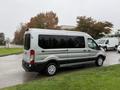 A silver 2019 Ford Transit van with large windows and a high roof parked on grass with wet pavement in the foreground