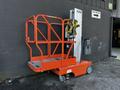 A 2000 JLG 12SP aerial lift with a bright orange platform and safety harness attached against a gray wall
