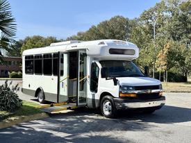 A 2014 Chevrolet Express bus with a white and gray exterior features an open doorway and accessibility ramp at the front