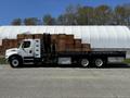 A white 2012 Freightliner M2 112 flatbed truck with a black bed and a mounted crane on its side, parked beside a stack of wooden crates