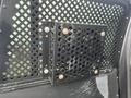 A black metal partition with a grid pattern designed to separate the front and back areas of a vehicle, featuring a square vent with rounded corners secured by bolts