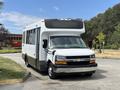 A 2020 Chevrolet Express bus in white with a black stripe on the bottom facing forward with a large front window and roof mounted air conditioning unit