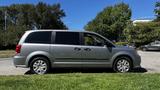 A silver 2016 Dodge Grand Caravan parked on a grassy area with a side view showing its sliding doors and five-spoke wheels