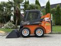 A 2017 Gehl R105 skid steer loader with a black and orange design and a front bucket is positioned sideways on a paved surface
