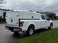 A white 2017 Ford F-150 with a cap on the truck bed parked on a street with grass on the side