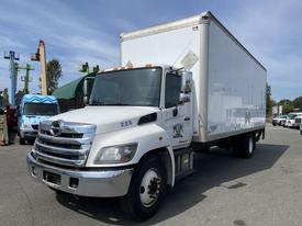 A 2018 Hino 338 box truck with a white body and silver grille parked in a commercial area with its headlights on and orange markers visible on the roof