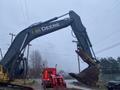 A 2014 John Deere 350G excavator with a large bucket raised and an orange-red truck in the background