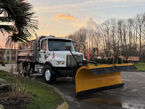 A 2006 Freightliner M2 106 truck with a yellow snow plow attached at the front and flags on the sides parked on a driveway