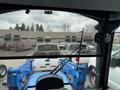 A 2016 New Holland T475 tractor with a blue body and loader attachments viewed from the inside of a cab looking out towards other vehicles