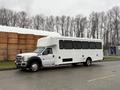 A white 2011 Ford F-550 bus with multiple windows and a large boxy shape parked on a road with trees in the background