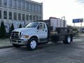 A white 2011 Ford F-750 truck with a flatbed and a large tank on the back parked on a street