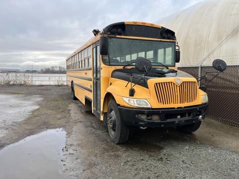 A 2009 International 3000 school bus painted yellow with a black front and side mirrors parked on gravel with a cloudy sky in the background